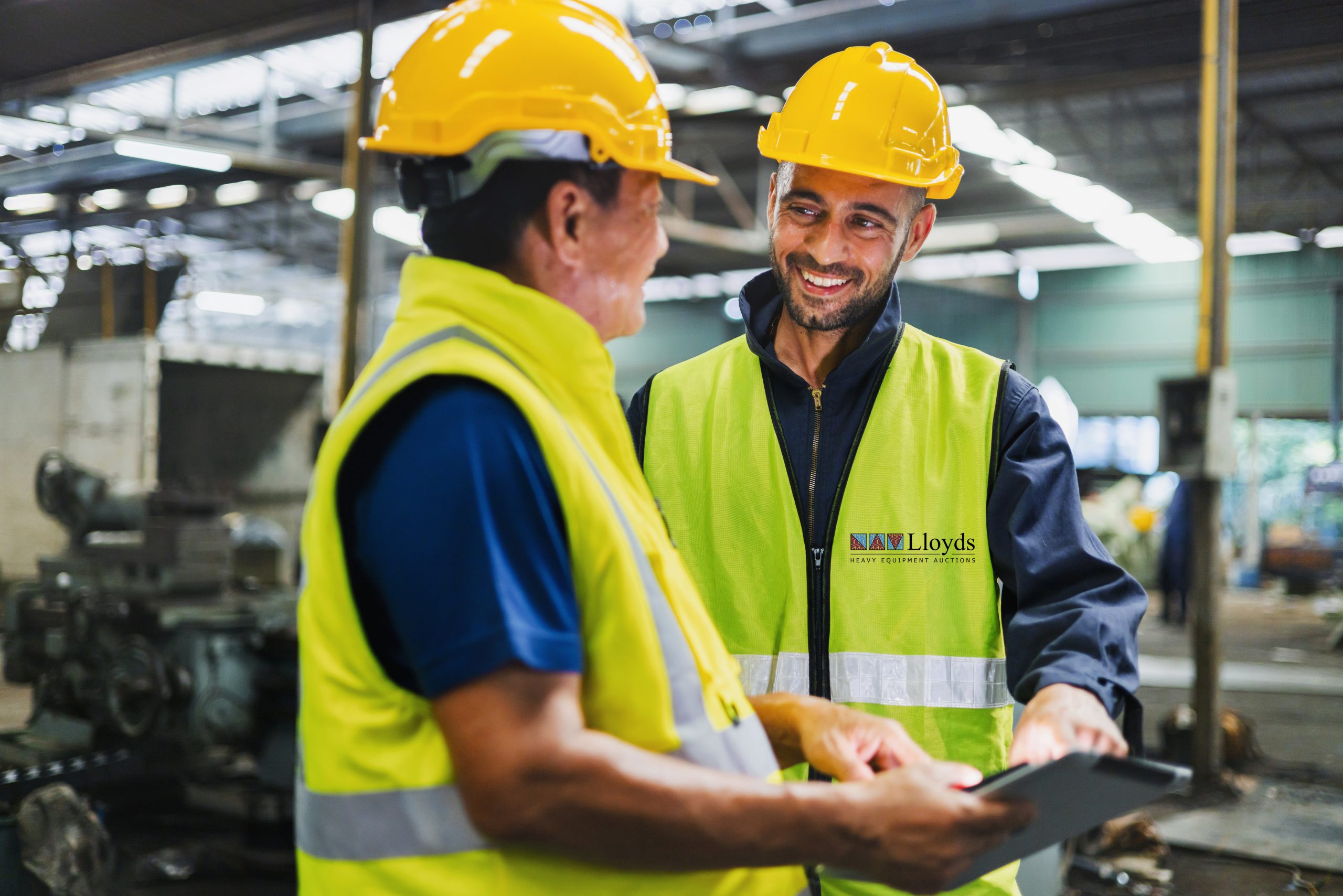 Two men in safety gear are talking to each other. One of them is holding a tablet. Scene is friendly and professional